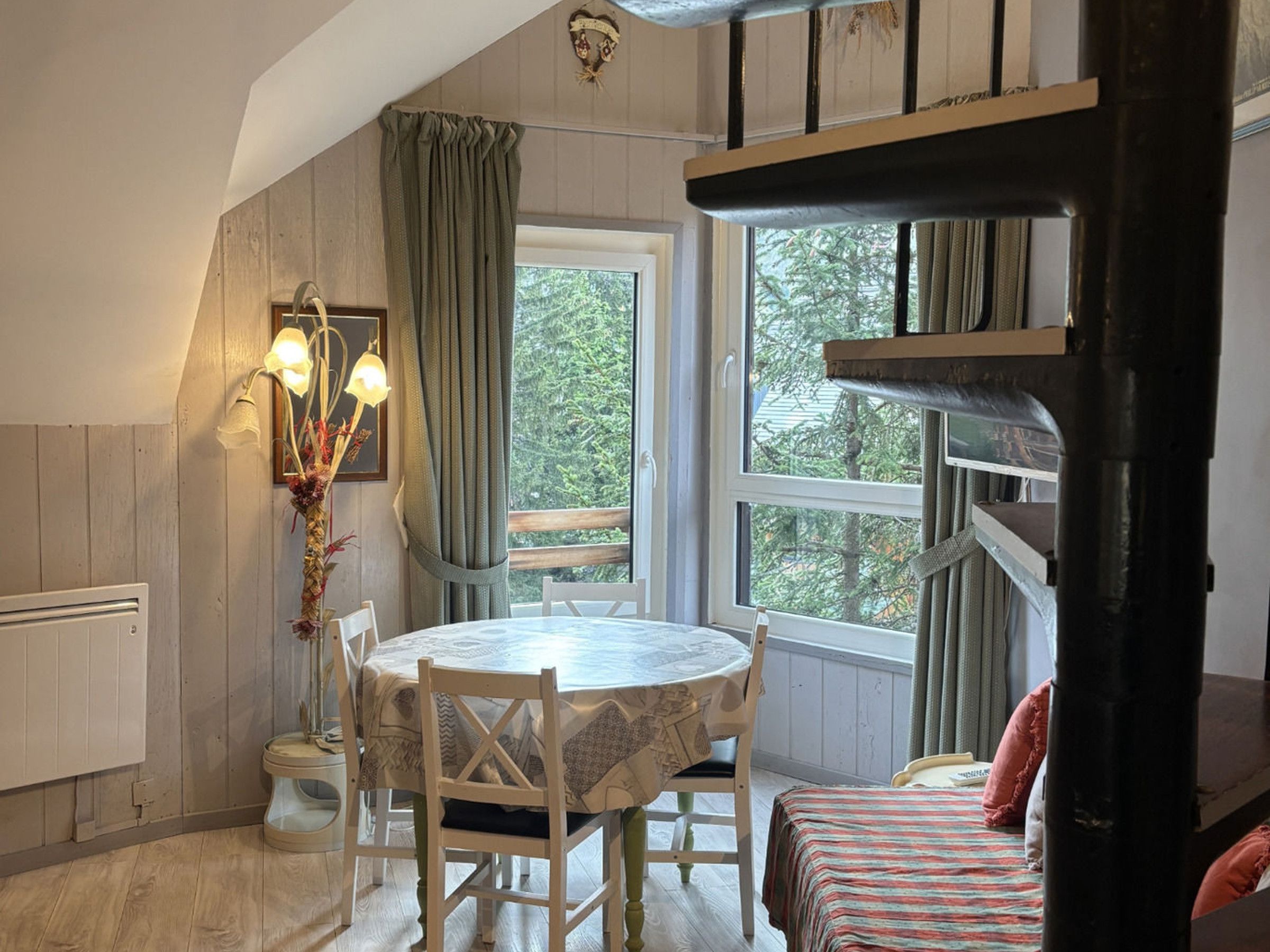 Quarto aconchegante com mesa redonda, cadeiras, janela com cortinas, radiador e beliche com almofadas.