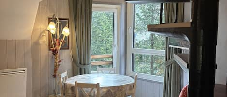 Quarto aconchegante com mesa redonda, cadeiras, janela com cortinas, radiador e beliche com almofadas.