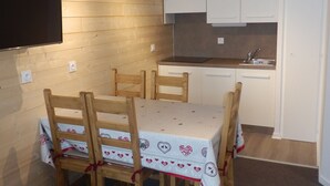 Eine kompakte Küche mit Mikrowelle, Spüle und weißen Schränken. Ein Esstisch mit Stühlen sowie ein an der Wand befestigter Fernseher sind vorhanden.