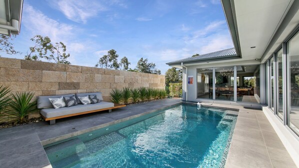 A modern outdoor pool area with a stone wall, a built-in bench, and a glass-enclosed dining area. - HUNTER VALLEY WILDFLOWER (POKOLBIN)