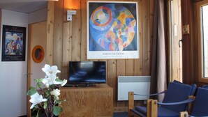 Een kamer met houten paneelwanden, een lijst met een abstract schilderij, een televisie, een witte radiator en een blauwe stoel.