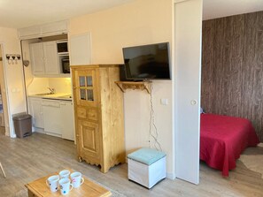 Une pièce compacte comprenant un coin cuisine, un lit recouvert d’une couverture rouge, un téléviseur fixé au mur et une petite table avec des tasses.