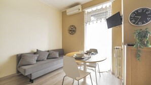 A living room with a sofa, a dining table with chairs, a TV mounted on the wall, and a clock. - GuestHost - Studio with terrace 100m from M1 subway red line! (Milano)