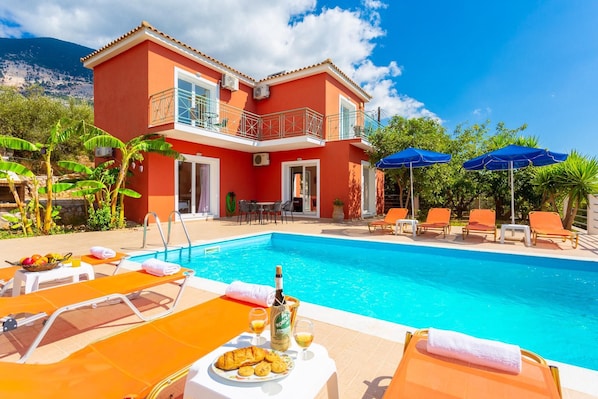 A two-story building with a swimming pool, outdoor seating, and a mountain backdrop. - Villa Georgia in Vlachata (Kefalonia)