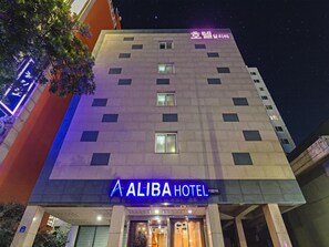 Un hôtel avec une enseigne indiquant « HÔTEL ALIBA » et un panneau lumineux au néon au-dessus.