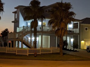 A multi-story building with balconies, a white picket fence, and palm trees. - 3 Story Home with Elevator, Walk to Beach, Shared Pool and Golf Cart Included (Port Aransas)