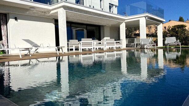 Een modern hotel met een zwembad, witte lounge fauteuils en een helderblauwe lucht.