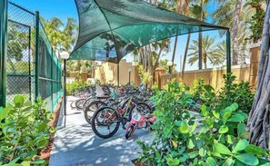 Bicycles are parked under a shaded canopy surrounded by lush greenery and palm trees.