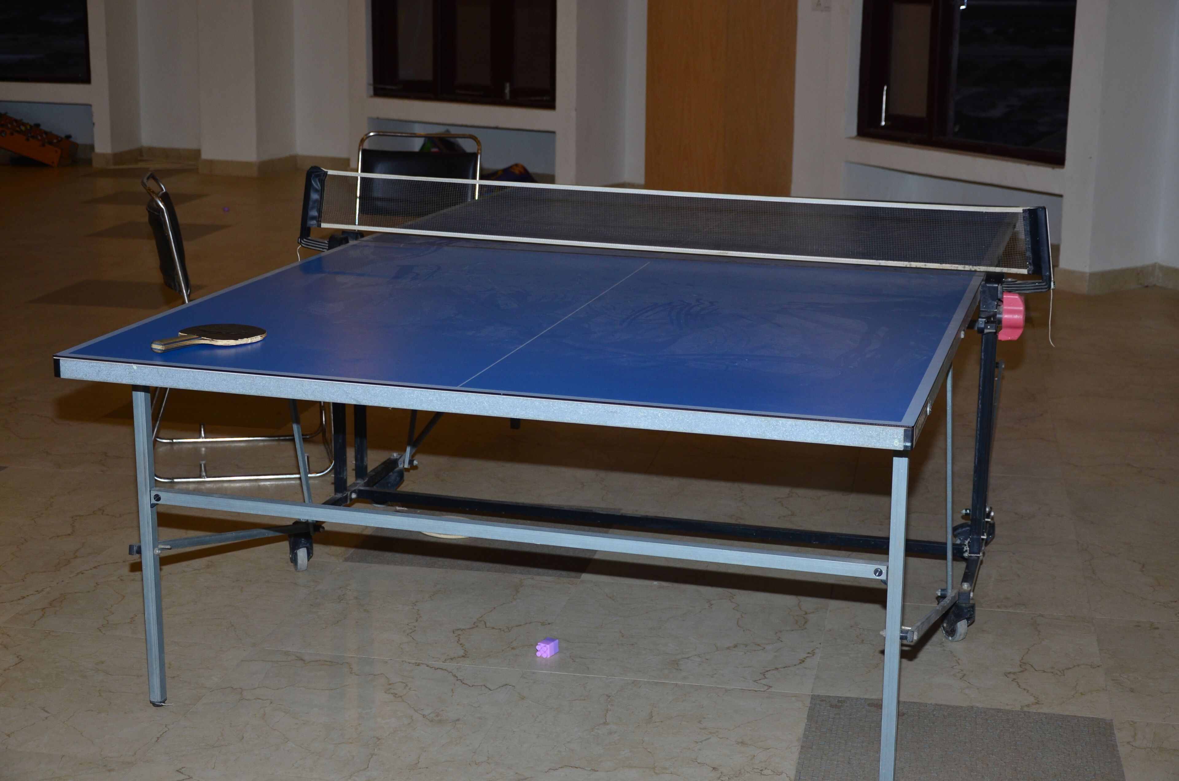 Une table de ping-pong avec une raquette, une chaise et une table sur roulettes, dans une pièce dotée de fenêtres et d’un sol carrelé.