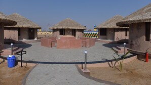 A courtyard with thatched-roof huts, a central seating area, and a blue trash bin. - Praveg White Rann Resort Rann Utsav Dhordo (Bhuj)