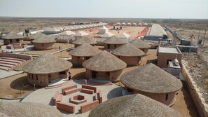 A desert resort with thatched-roof huts arranged in rows, a central courtyard, and a long building with a white roof.