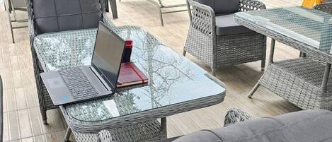 Un espace aménagé au bord de la piscine, avec des meubles en osier, un ordinateur portable posé sur une table en verre, et une piscine en arrière-plan.