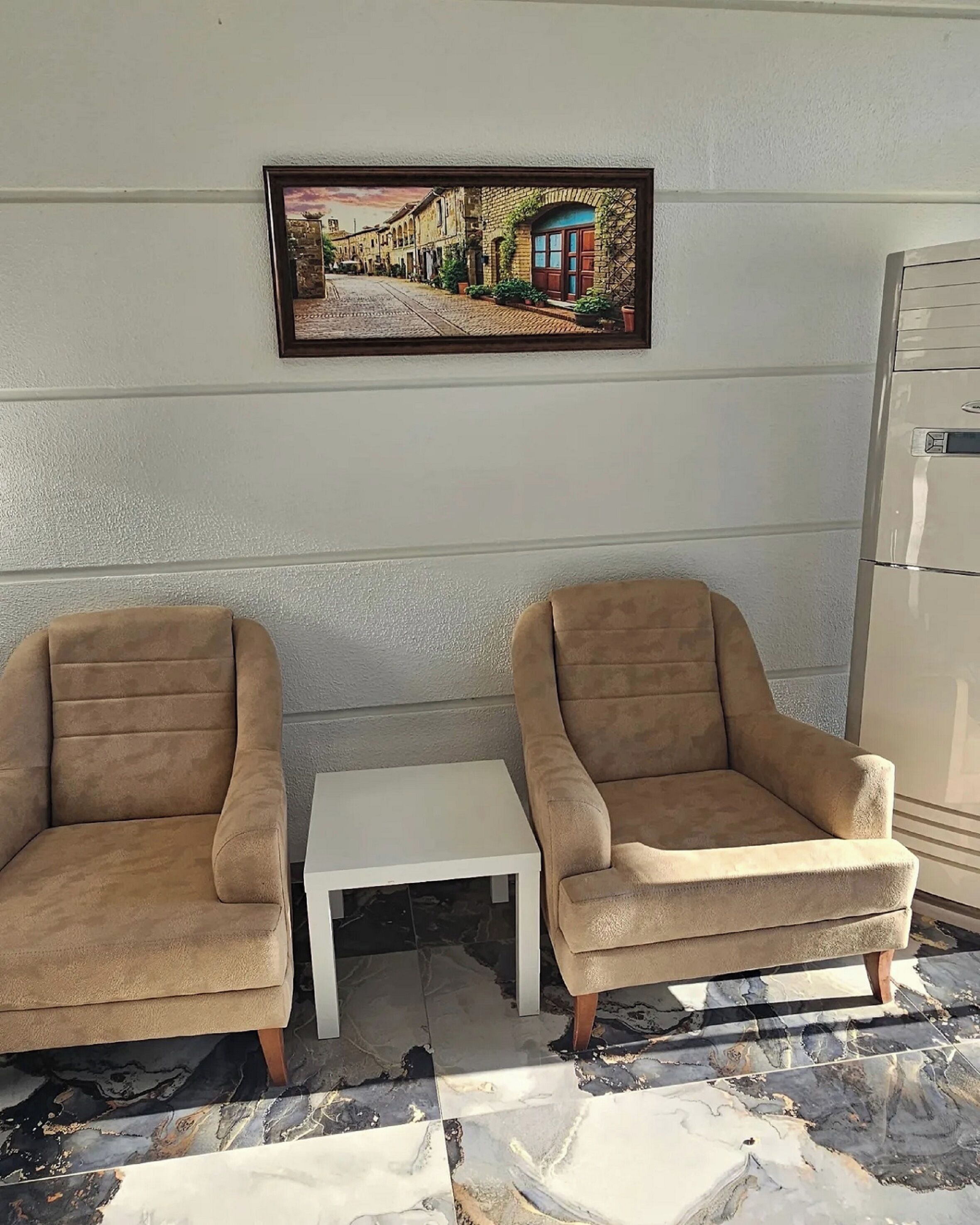 A room with two beige armchairs, a small white table, and a framed picture of a street scene on the wall.