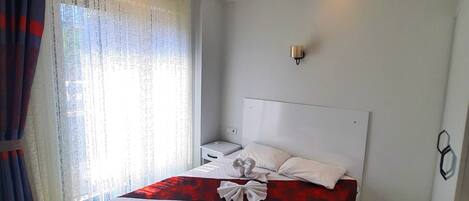 A neatly made bed with a white headboard and a red accent, a bedside table with a lamp, and a window with sheer curtains.