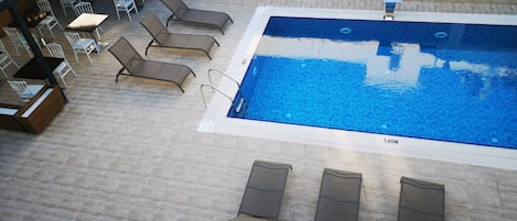 An indoor swimming pool with lounge chairs and tables, surrounded by a tiled deck.