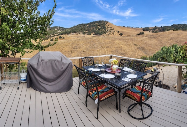 An outdoor deck with a covered grill, dining table, chairs, and a scenic view of hills.