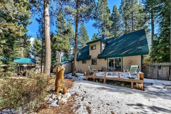 Una cabaña con terraza y zona de estar exterior, rodeada de altos árboles y nieve en el suelo.