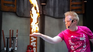 Eine Person tritt mit Feuer in einem Veranstaltungsraum auf, im Hintergrund eine Metallkonstruktion und ein Schild.