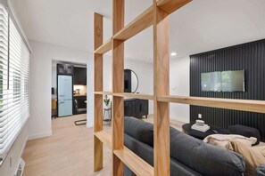 A modern living room with a grey sofa, wooden shelves, a TV mounted on a dark wall, and a kitchen in the background. - Newly Updated 1 BR Flat Near Downtown A2 (Ann Arbor)