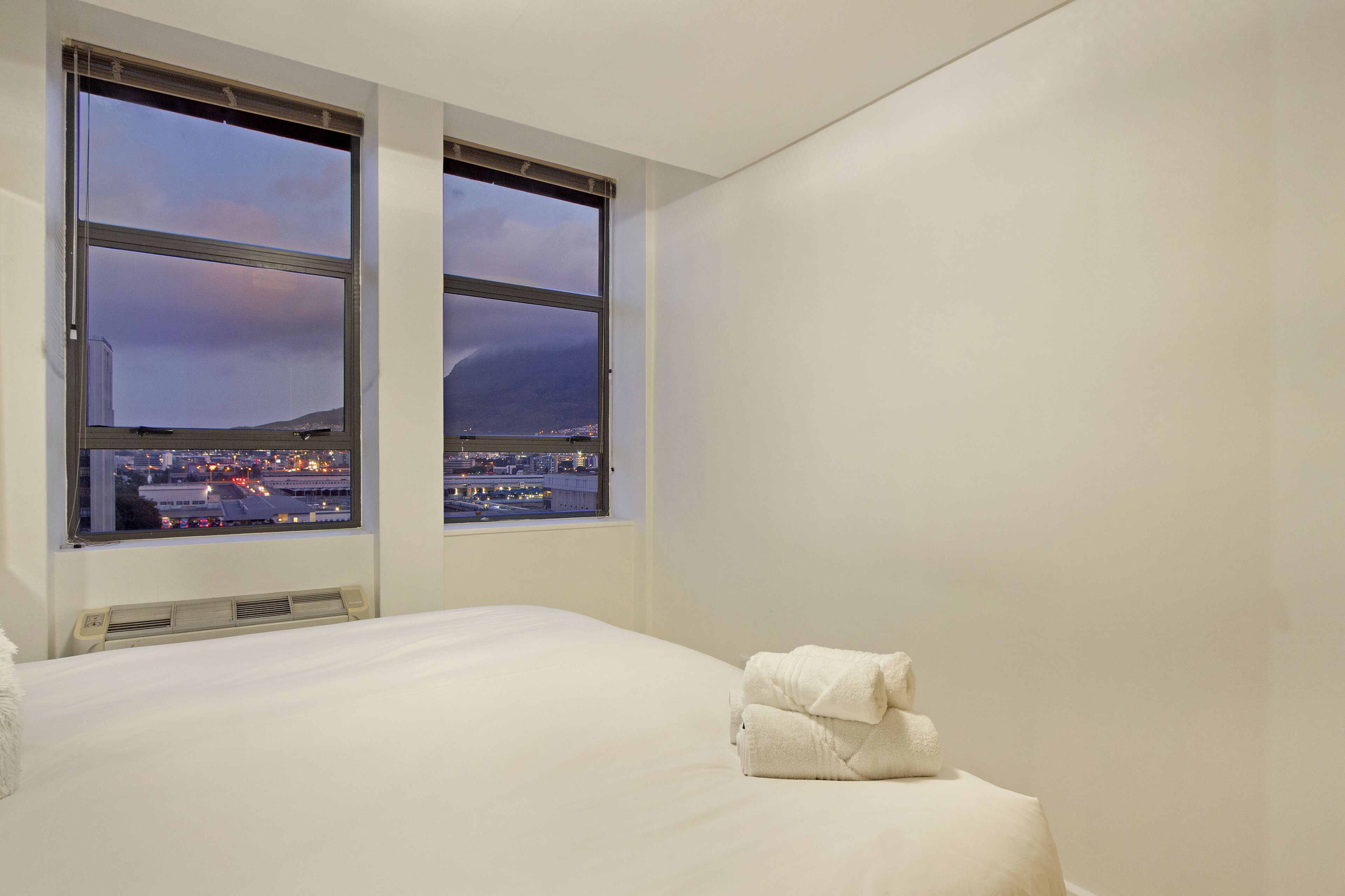 A hotel room with a large bed, two windows with a city view, and a stack of towels on the bed.