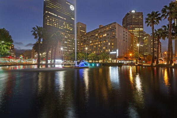 A nighttime cityscape with high-rise buildings, a calm water body reflecting lights, and palm trees lining the area. - Fountain Suite 906 (Cape Town)