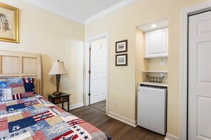 Quarto com colcha acolchoada, luminária de cabeceira, mesa pequena, geladeira e porta que dá para outro cômodo.