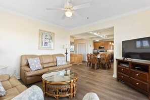 A living room with a sofa, a glass-top coffee table, wicker chairs, a dining table with chairs, a kitchen area, and a TV on a wooden stand. - Oceanview 4BR/4BA Condo, Steps to Beach/Pool (Ocean Isle Beach)