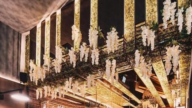 Ein Restaurant mit Barbereich, Tischen, die mit Tellern und Gläsern gedeckt sind, und hängenden Blumendekorationen.