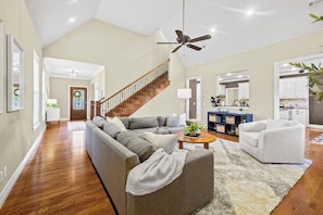 A spacious living room with a wooden staircase, a grey sectional sofa, a white armchair, and a coffee table. - Willow Tree Retreat-POOL-10 min to U of A (Fayetteville)