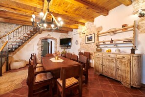 A rustic dining area with a wooden table, chairs, and a chandelier. A staircase with a wooden railing leads to an upper level.