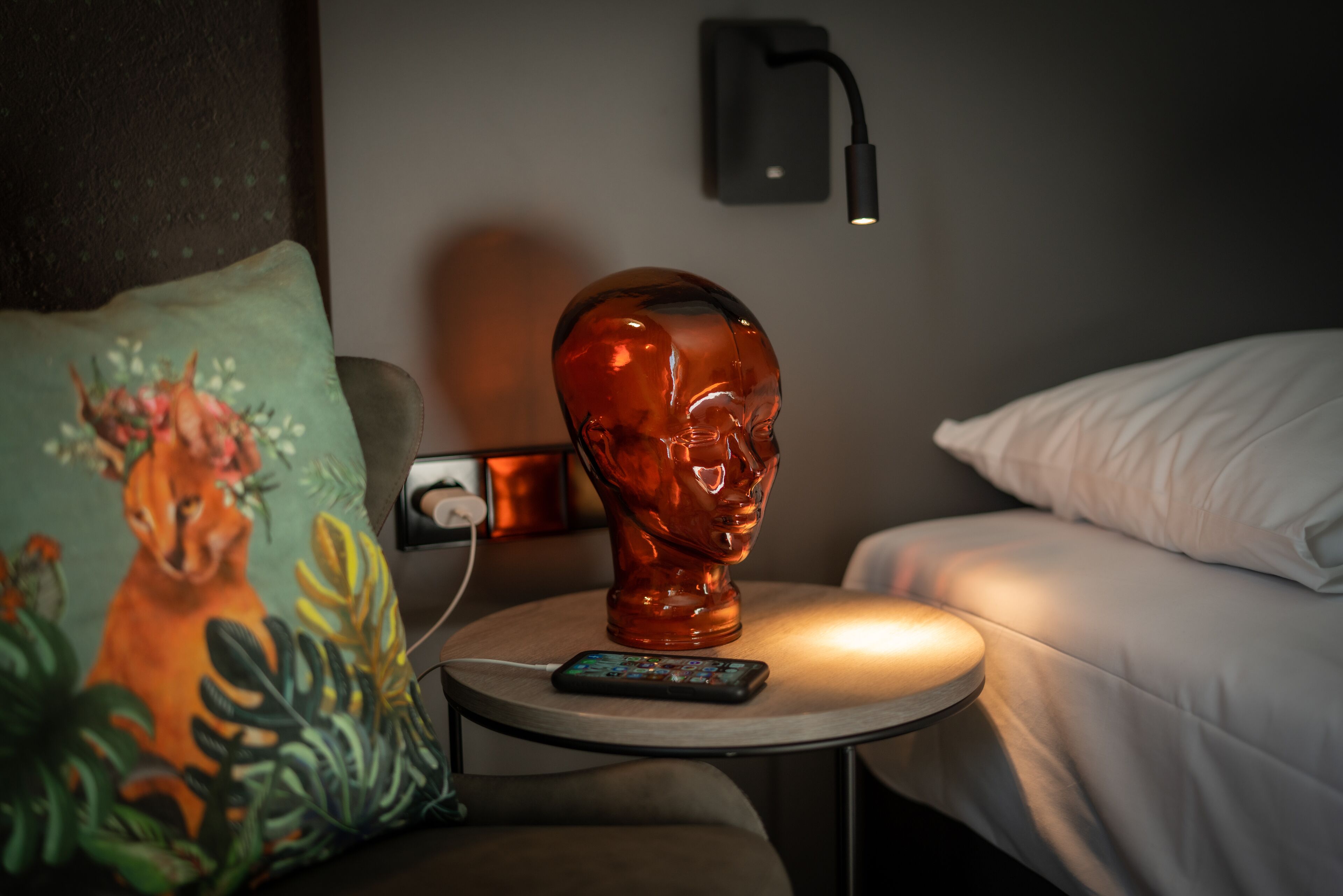 Una habitación con cama, una mesita de noche con lámpara, una escultura decorativa en la cabecera y un almohadón con diseño floral.