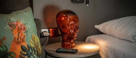 Una habitación con cama, una mesita de noche con lámpara, una escultura decorativa en la cabecera y un almohadón con diseño floral.
