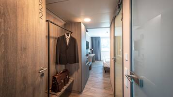 A hotel corridor with a wooden wall, a coat rack with a suit, a leather bag, and a bed in the distance.