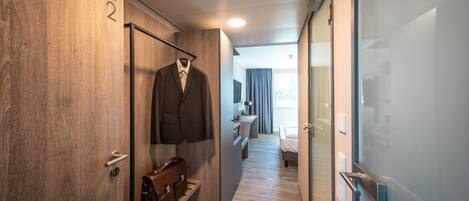 Un couloir d’hôtel avec un mur en bois, un porte-manteau sur lequel est posé un costume, un sac en cuir, et un lit visible au loin.