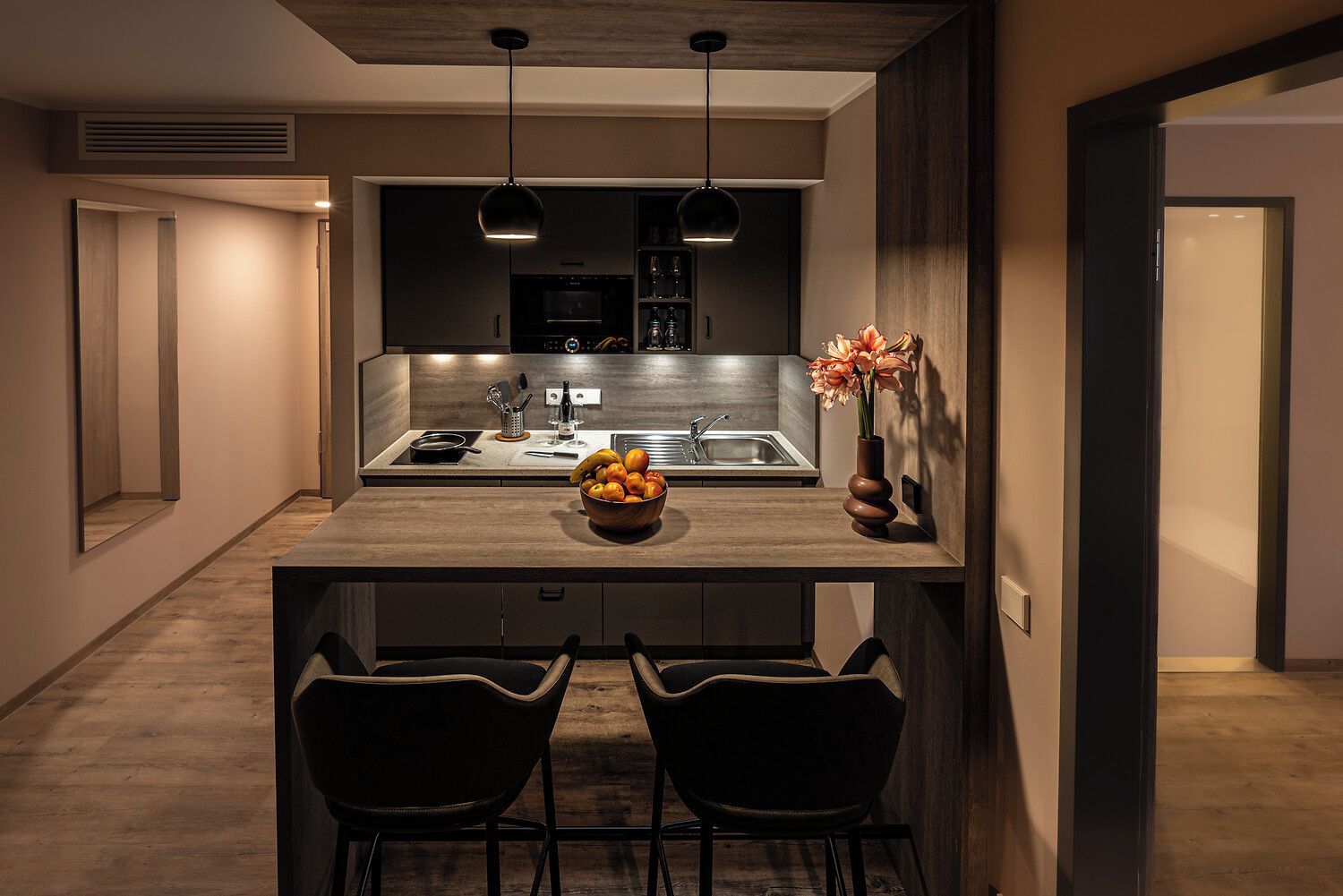A modern kitchen with a central island, two bar stools, pendant lighting, and a bowl of fruit on the island.