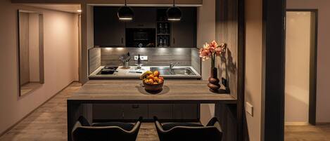 A modern kitchen with a central island, two bar stools, pendant lighting, and a bowl of fruit on the island.