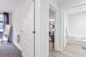 A hotel room with a white door, a view of the living area, and a bathroom with a white bathtub and sink.