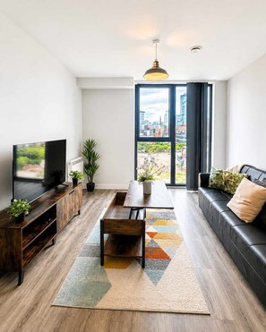 A modern living room with a flat-screen TV, a wooden coffee table, a sofa, and a large window offering a city view.