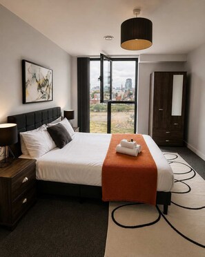 A modern hotel room with a large bed, a city view, a dark wooden wardrobe, and a painting on the wall.