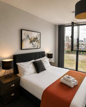A hotel room with a bed, two bedside tables, a city view, and a modern abstract painting on the wall.