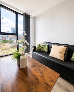 A modern living room with a black leather sofa, a wooden coffee table, and a large window offering a city view.