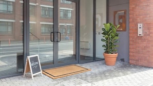 A modern building entrance with glass doors, a welcome sign, a potted plant, and a brick wall. - Stylish & Bright 1-Bedroom Apartment in Birmingham (Birmingham)