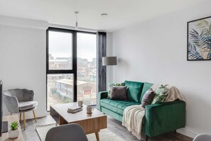 Een moderne woonkamer met een groene bank, een grijze fauteuil, een houten salontafel en een stadszicht door grote ramen.