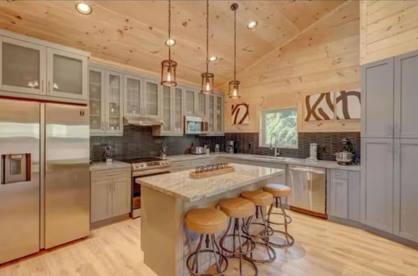 A modern kitchen with a central island, bar stools, stainless steel appliances, and wooden ceiling.
