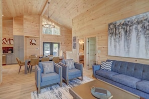 A spacious living area with a large sofa, two armchairs, and a coffee table. There is a dining area with a table and chairs, and a kitchen visible in the background.