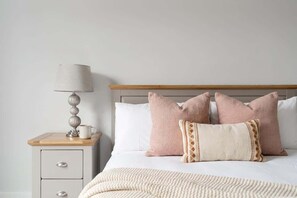 Una cama bien hecha con edredón blanco y varios almohadones, una lámpara de noche y una mesita de noche de madera con cajones.