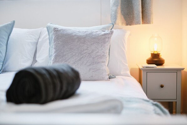 Ein ordentlich bezogenes Bett mit weißen und grauen Kissen, einer aufgerollten schwarzen Decke und einem Nachttisch mit einer beleuchteten Lampe.