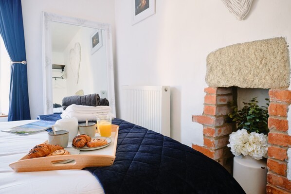 A neatly made bed with a dark blue quilt, a breakfast tray with croissants and orange juice, and a brick fireplace with white flowers. - Scallop Cottage (Porthleven)