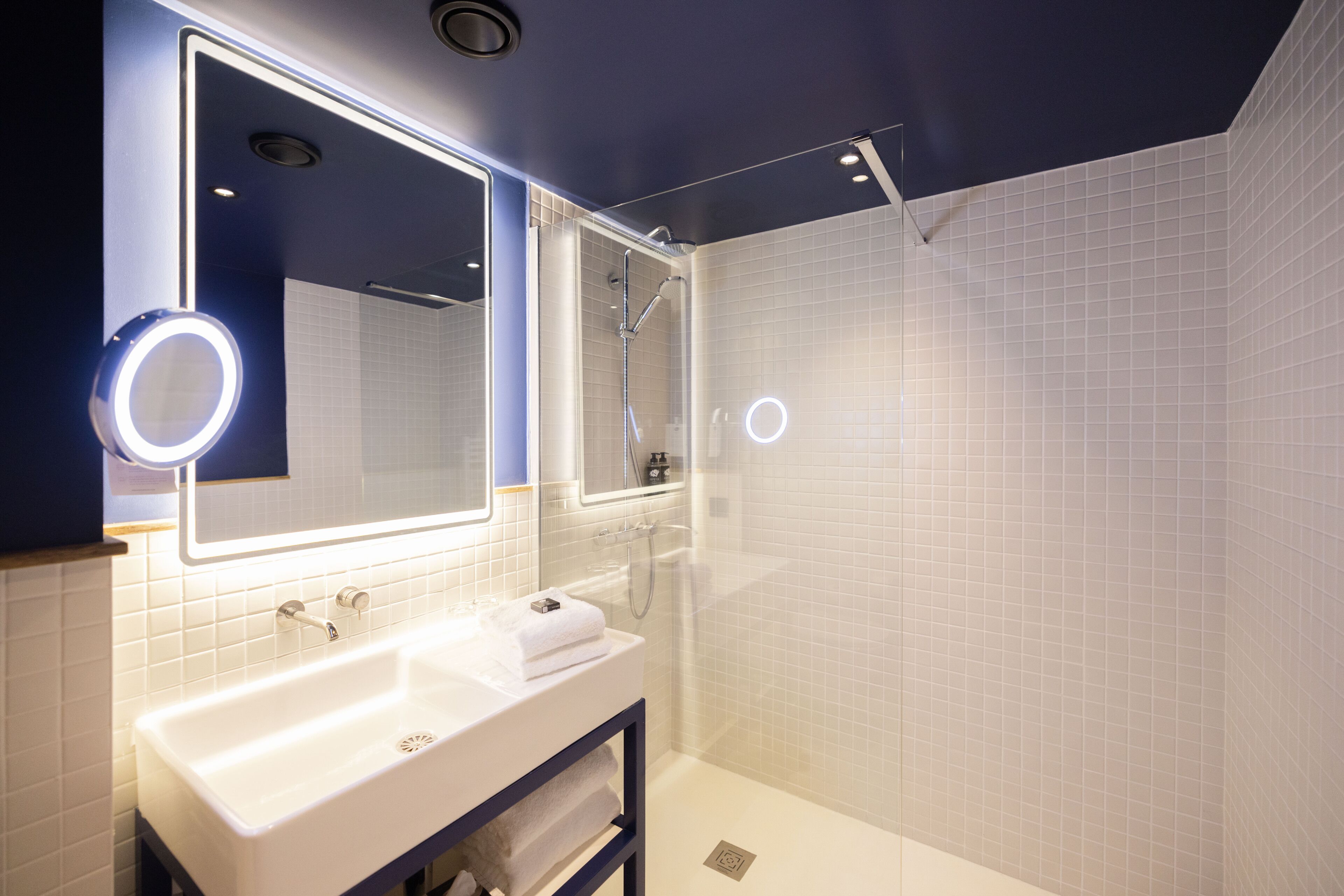 A modern bathroom with a white sink, a large mirror with a circular light, and a glass-enclosed shower.