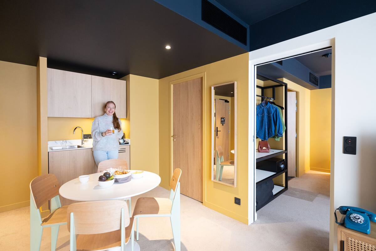 A modern kitchen with a woman preparing food, a dining area with a table and chairs, and a walk-in closet with hanging clothes.
